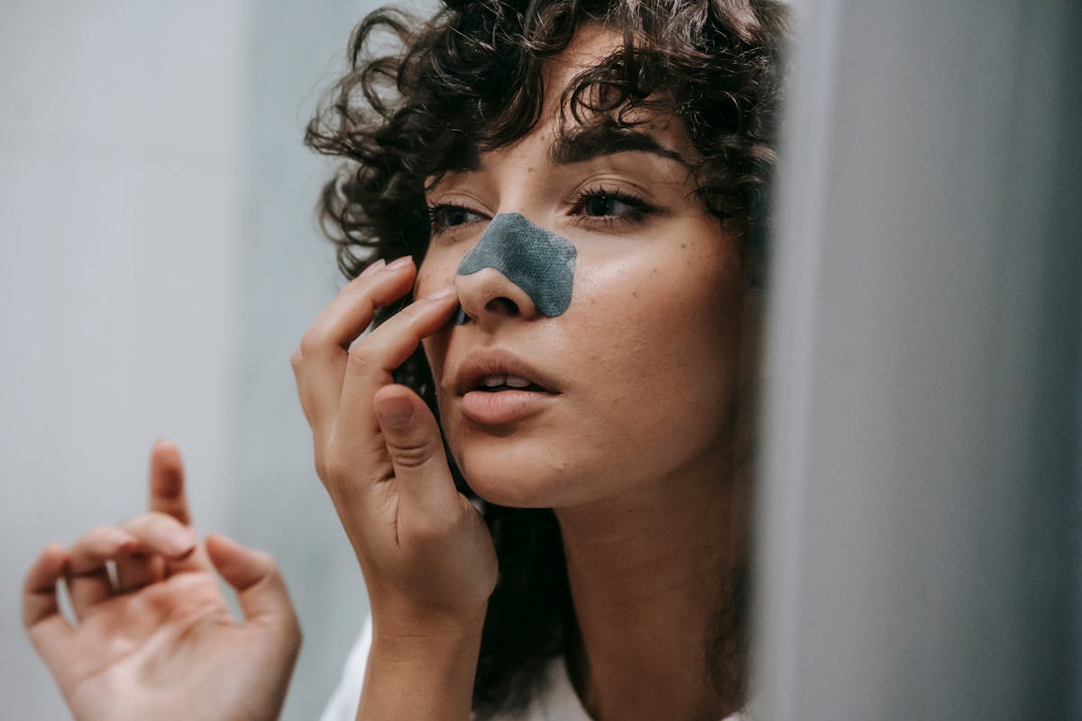 Photo by Sam Lion Mirror reflection of young female with curly hair putting nose strip mask on face in bathroom
