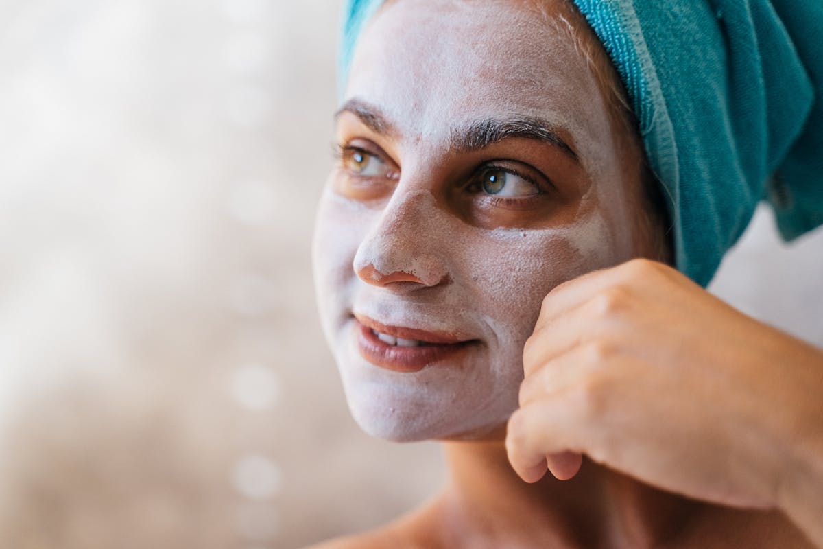 Smiling woman with a facial mask and towel for skincare indoors.
