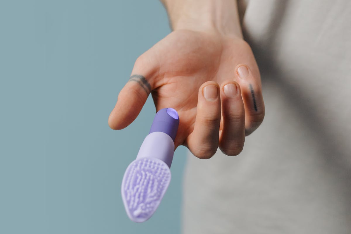 A close-up image showing a hand holding a purple silicone finger vibrator against a neutral background.