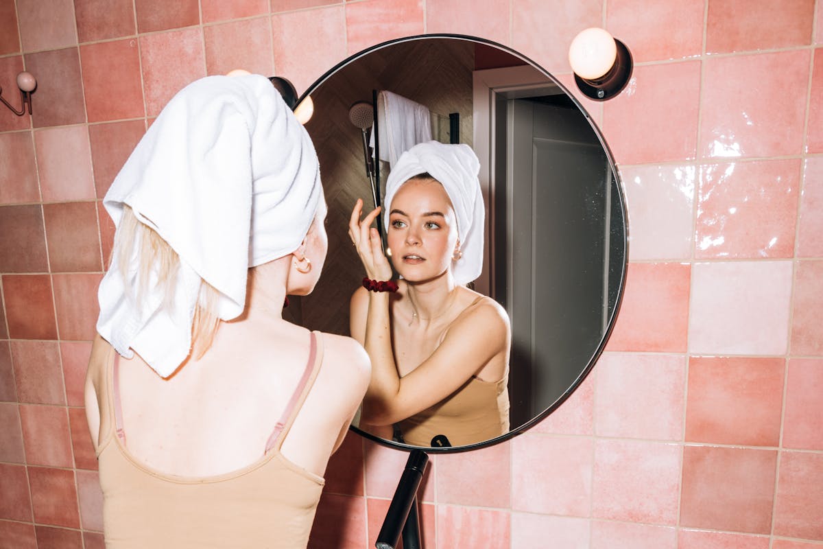 A young woman with a towel in a pink bathroom, reflecting in round mirror as she performs skincare.