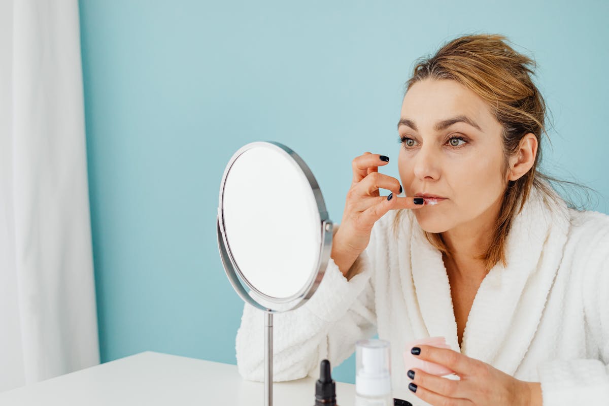 Photo by Karola G Caucasian woman in a bathrobe applies skincare in front of the mirror against a pastel blue background.