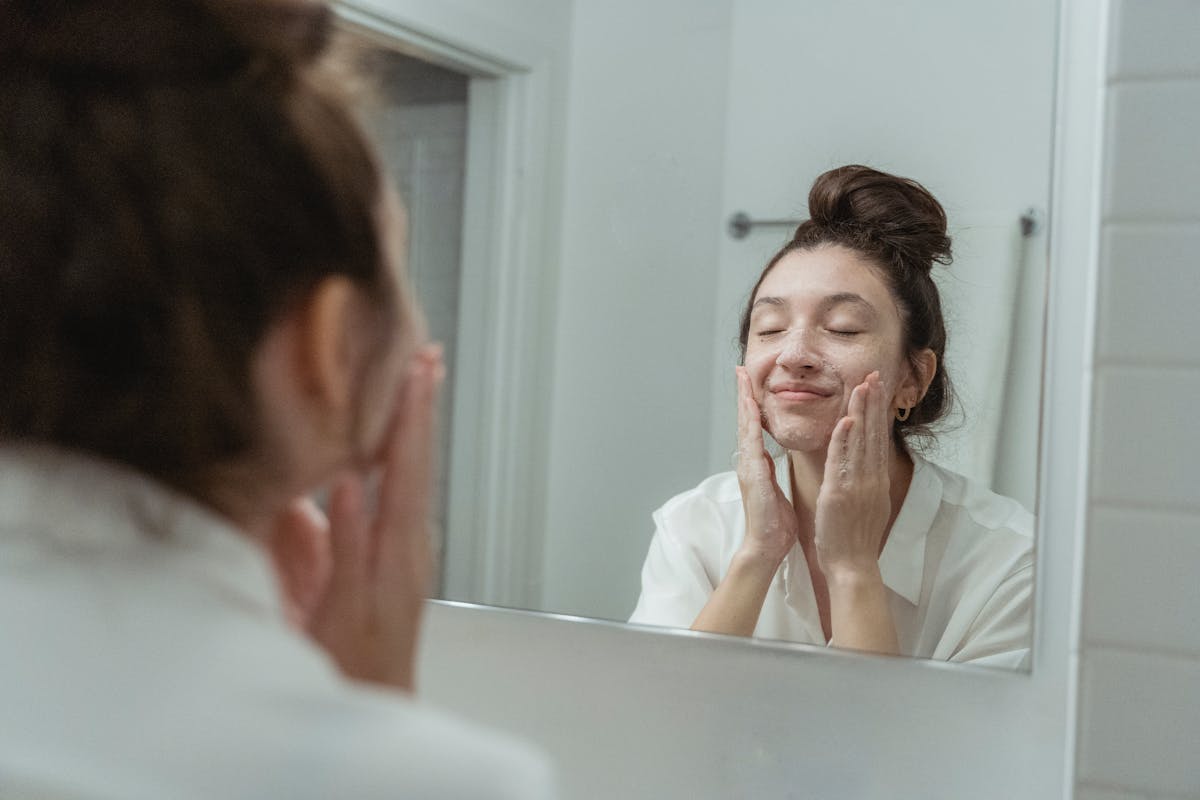 Photo by Miriam Alonso Woman applying soap to her face, reflected in the bathroom mirror, enjoying a skincare routine.