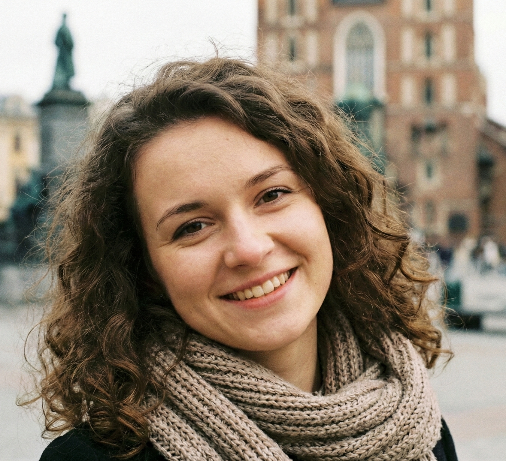 Młoda kobieta z kręconymi włosami uśmiecha się na tle historycznej architektury, nosząc beżowy szalik.