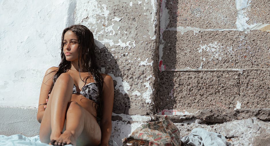 cover-image-276697 Młoda kobieta w kostiumie kąpielowym siedzi na plaży, opierając się o ścianę. Jej długie, mokre włosy opadają na ramiona, a ona patrzy w dal, relaksując się w słonecznym otoczeniu. W tle widoczna jest tekstura ściany.