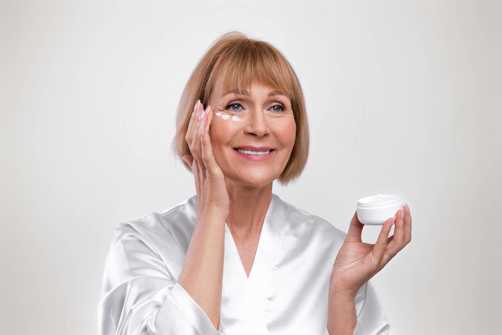 Kobieta w szlafroku nakłada krem na twarz, uśmiechając się. Trzyma w dłoni słoiczek z kosmetykiem, co sugeruje pielęgnację skóry i dbanie o urodę. Tło jest neutralne, co podkreśla jej naturalne piękno.